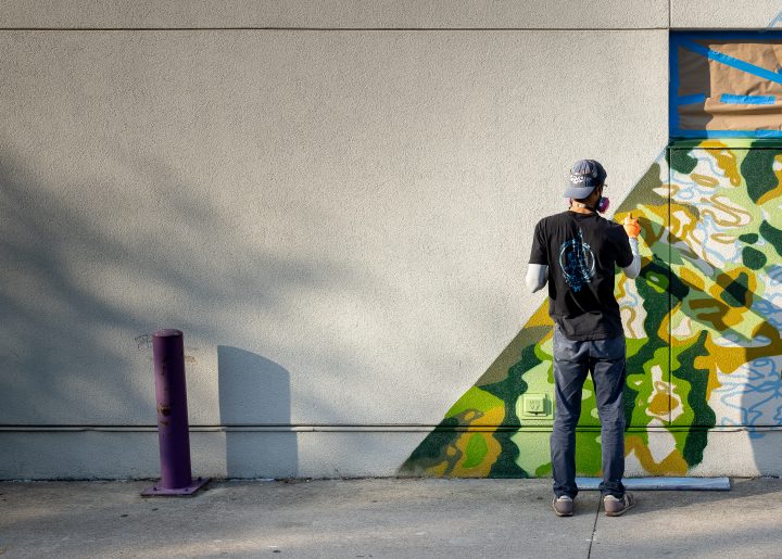 Muralist J Muzacz standing in front of his Fairtrade Month mural at Wheatsville in Austin, TX.