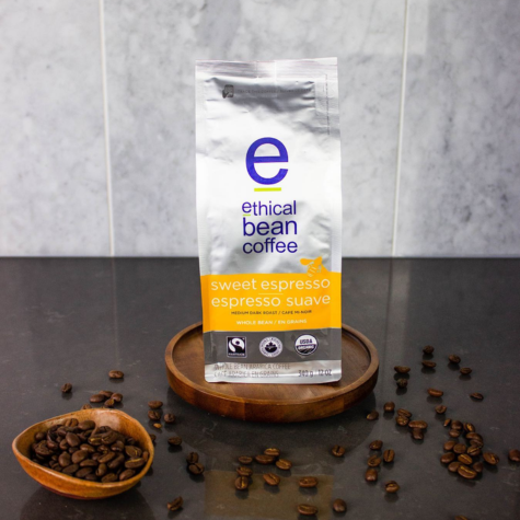 A package of sweet espresso whole bean Ethical Bean Coffee rests upon a wooden plate and is surrounded by coffee beans. The package includes both the Fairtrade Mark and USA organic labels.
