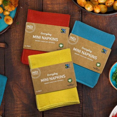 Packages of red, yellow and blue napkins rest upon a wooden table surrounded by food. The package labels identify the products as Everyday Mini Napkins, a part of the Indi By Kishu Line. The package label includes the Fairtrade Cotton Mark.