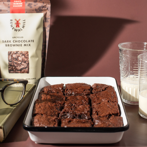 A tray of brownies rests on a counter amid a scene of coziness: glasses of milk, eyeglasses set on top of a book, and in the background, a package of Renewal Mill’s Upcycled Dark Chocolate Brownie Mix.