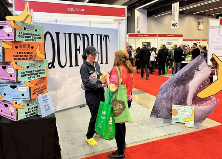 Equifruit booth at a convention, featuring boxes of bananas, two people in conversation, and a carboard cutout of a shark eating a banana.