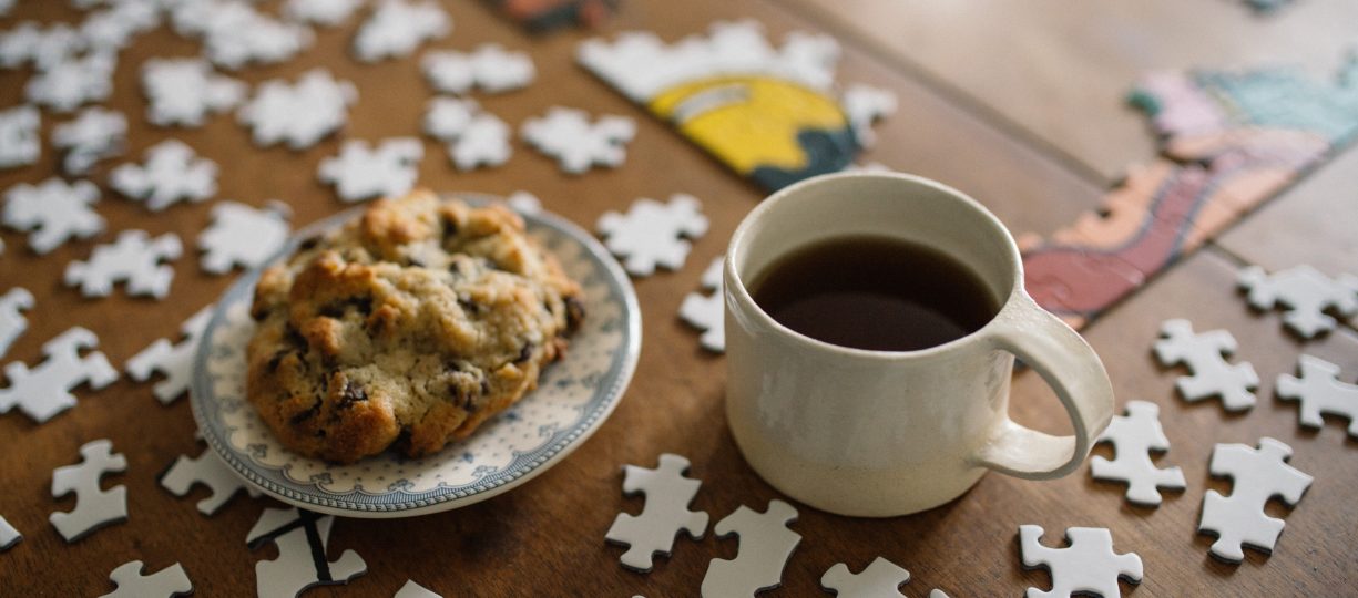 A jigsaw puzzle in the midst of coming together on a wooden table is accompanied by a scone and a cup of coffee.