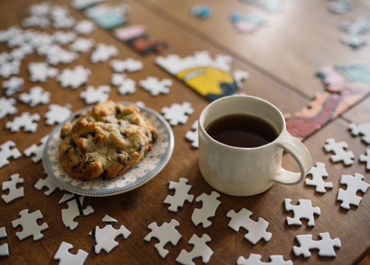 A jigsaw puzzle in the midst of coming together on a wooden table is accompanied by a scone and a cup of coffee.