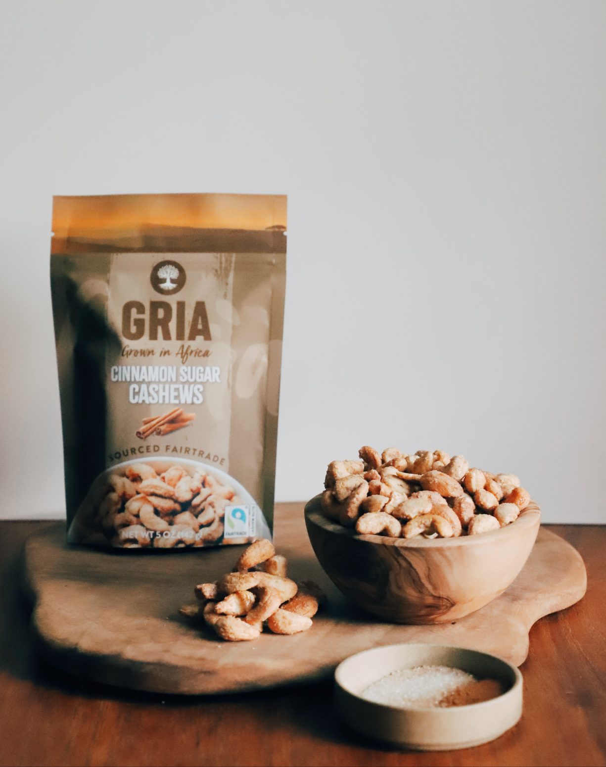 A bag of GRIA’s Fairtrade Cinnamon Sugar Cashews sits atop a wooden board. To the right of the bag is a wooden bowl displaying those cashews. Alongside it is a shallow bowl with cinnamon sugar.