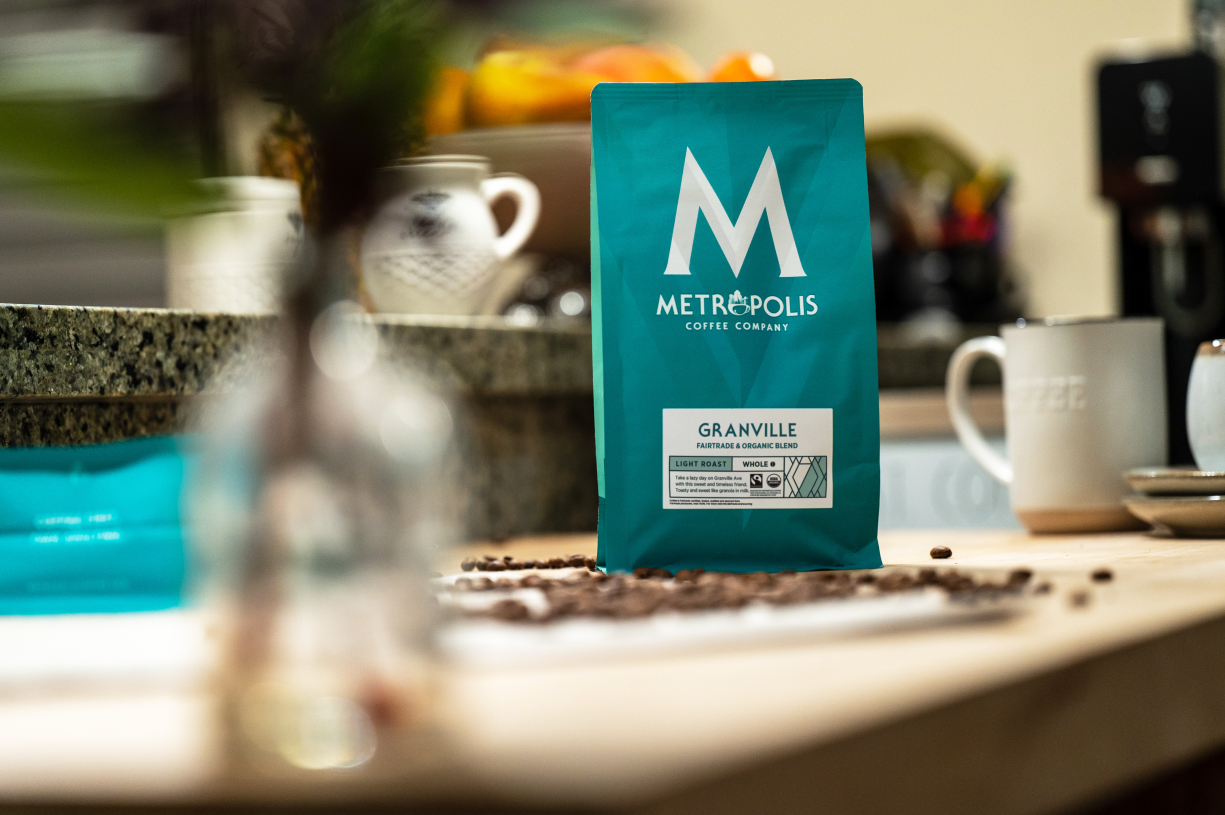 A bag of Metropolis Coffee Company's Granville, Fairtrade, Organic Coffee amid a wooden counter with coffee beans, a mug, and other kitchen items blurred in the background.