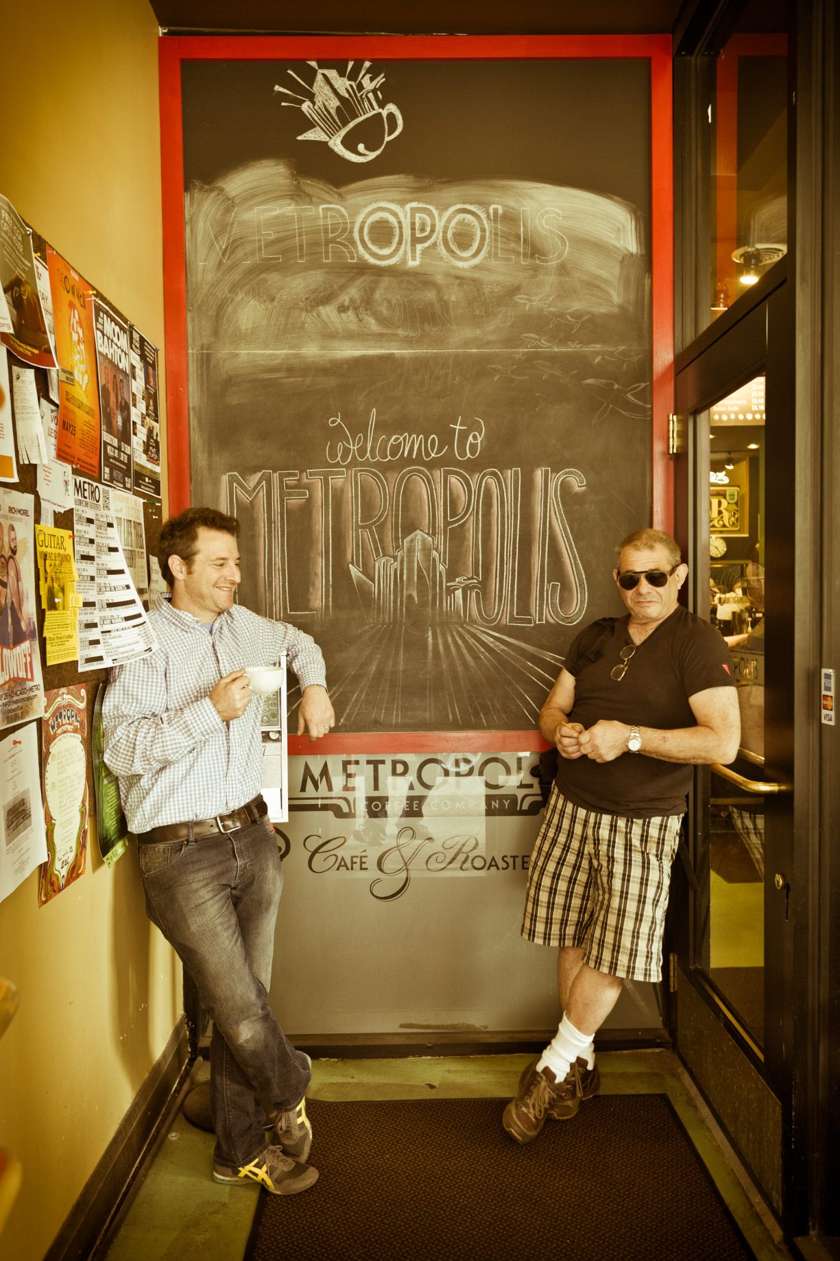 Tony and Jeff Dreyfuss lean against opposite walls in front a chalkboard that reads "Welcome to Metropolis." On left, Tony holds a cup of coffee in his hands. The photo is sepia-toned.