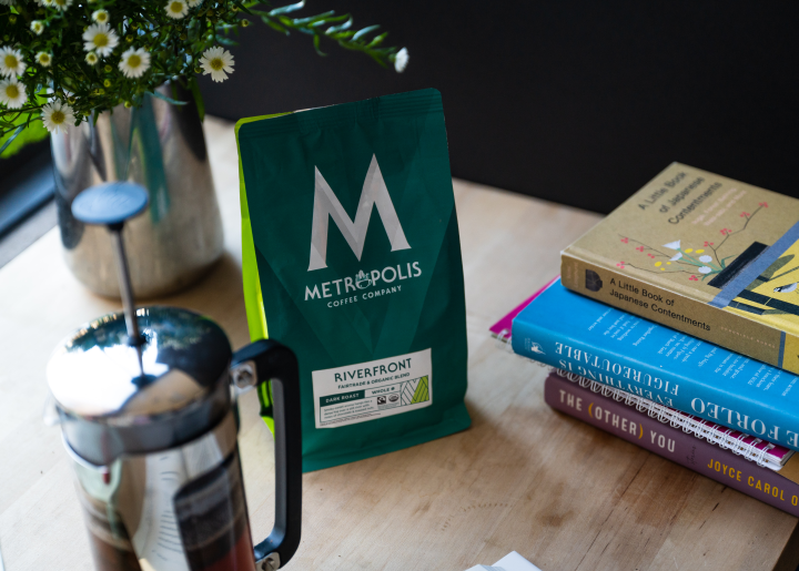 A wooden coffee table holds a vase of white asters, a French press, a bag of Metropolis Coffee Company's Riverfront Fairtrade, Organic Coffee, and a stack of books.